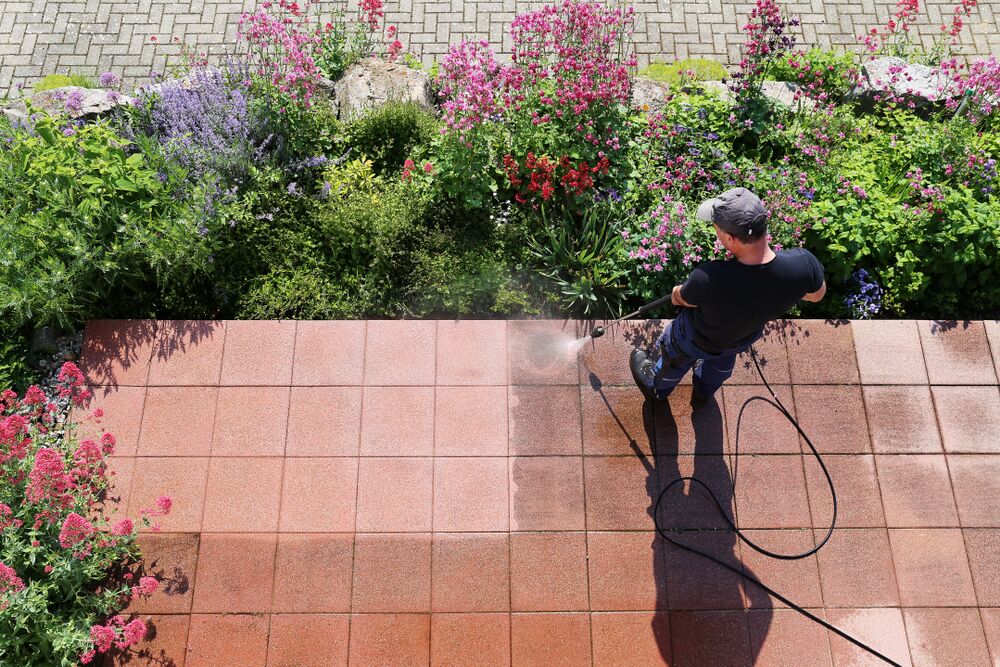 person pressure washing a red tile patio near a garden full of flowers